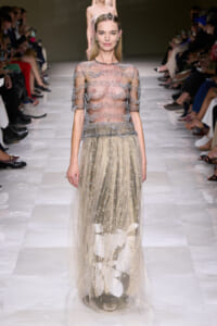 Model on a fashion runway wearing a sheer, beaded top with a glittering, floor-length tulle skirt featuring white leaf appliqués at the hem.