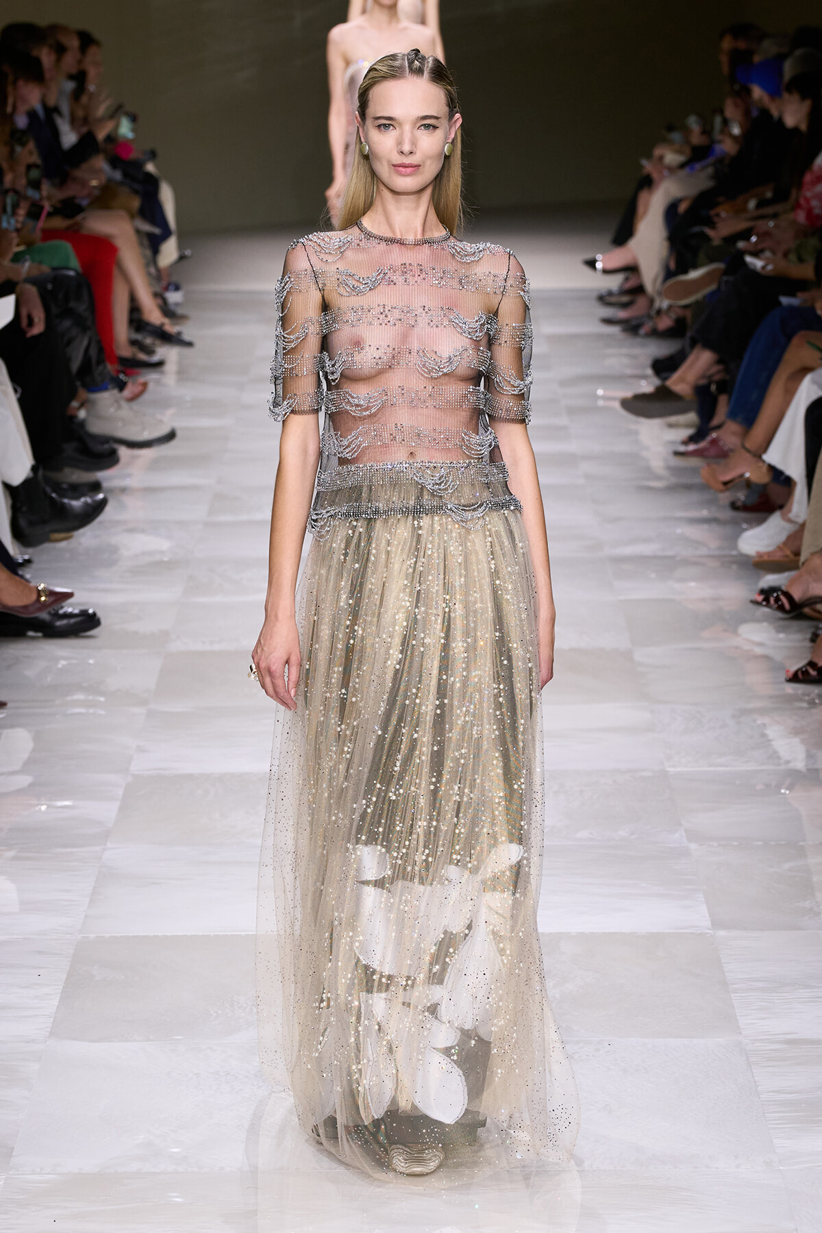 Model on a fashion runway wearing a sheer, beaded top with a glittering, floor-length tulle skirt featuring white leaf appliqués at the hem.