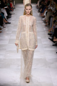 Model on a fashion runway wearing a sheer ivory gown heavily embellished with pearls, carrying a pearl-studded clutch.