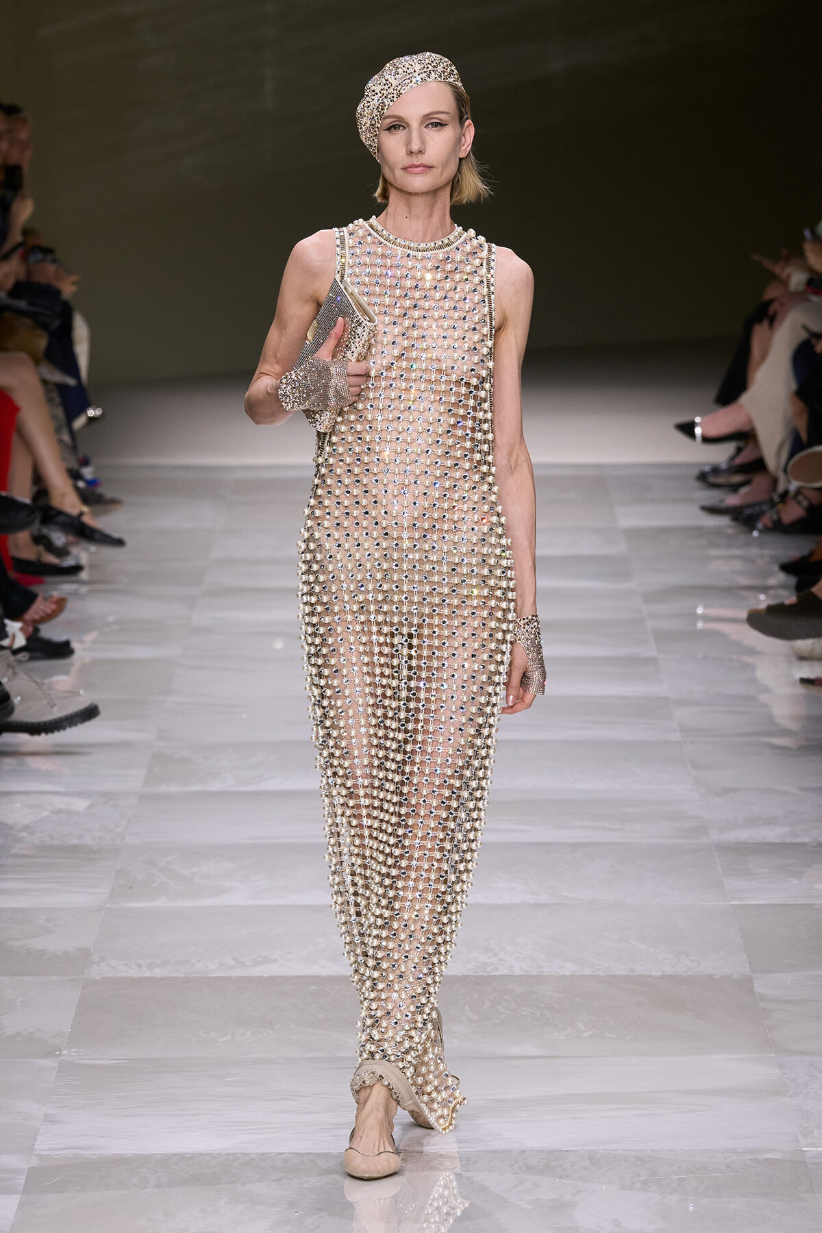 Model walks a runway in a nude, pearl-covered mesh dress with a matching beaded headscarf and gloves.