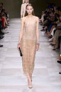 Model walking a fashion runway in a nude-beige beaded fringe dress, holding a black clutch along a glossy white catwalk.