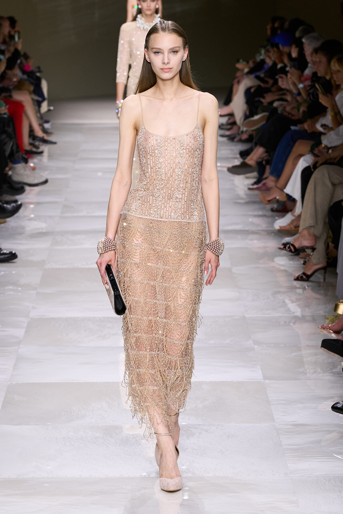 Model walking a fashion runway in a nude-beige beaded fringe dress, holding a black clutch along a glossy white catwalk.