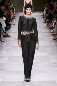 Model walking a runway in a black, sequined two-piece outfit with a cropped top and wide-leg pants, embellished with silver beads along the hem and cuffs, and a black beaded cap.