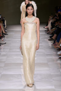 Model walks the runway in a strapless champagne sequined gown with a jeweled collar and pearl trim, clutch in hand.