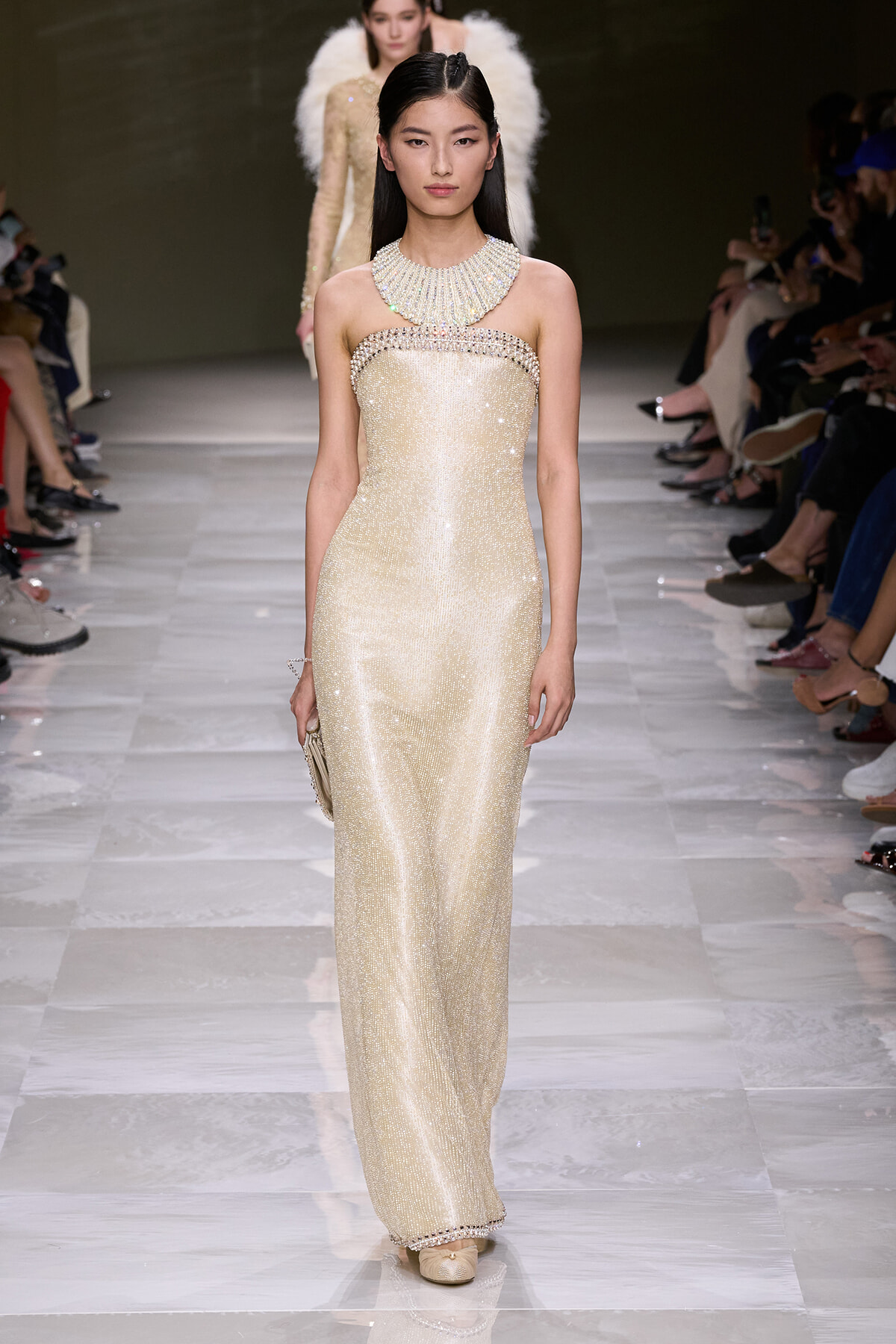 Model walks the runway in a strapless champagne sequined gown with a jeweled collar and pearl trim, clutch in hand.