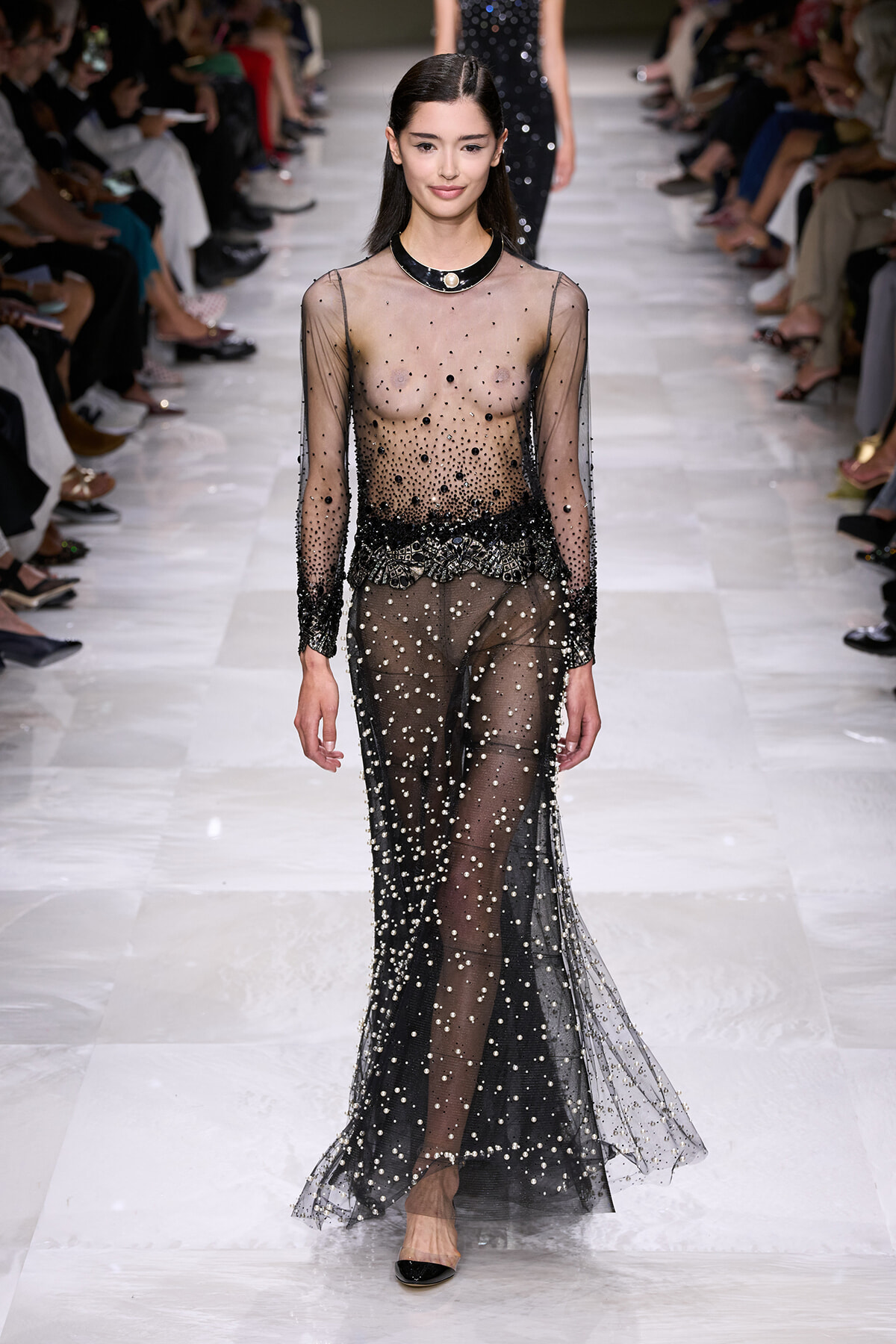 Model walking a fashion runway in a sheer black gown covered in pearls and beads, with an audience on both sides watching.