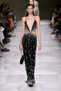 Model walks the runway in a black beaded gown with a deep V-neck and cutouts at the waist, carrying a small black clutch.