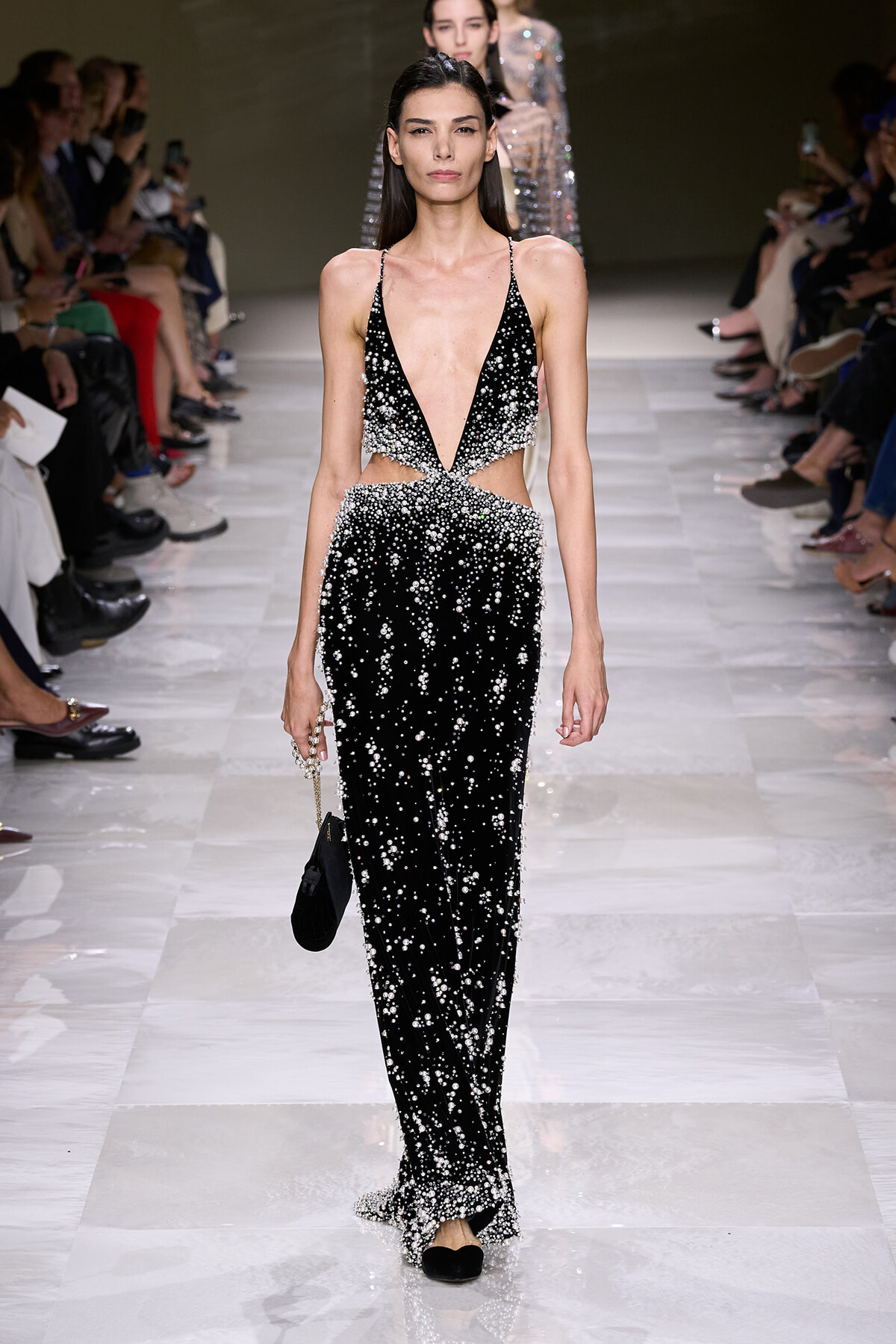 Model walks the runway in a black beaded gown with a deep V-neck and cutouts at the waist, carrying a small black clutch.