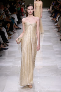 Model on a runway wearing a strapless champagne gown adorned with pearls, holding a small beaded clutch.