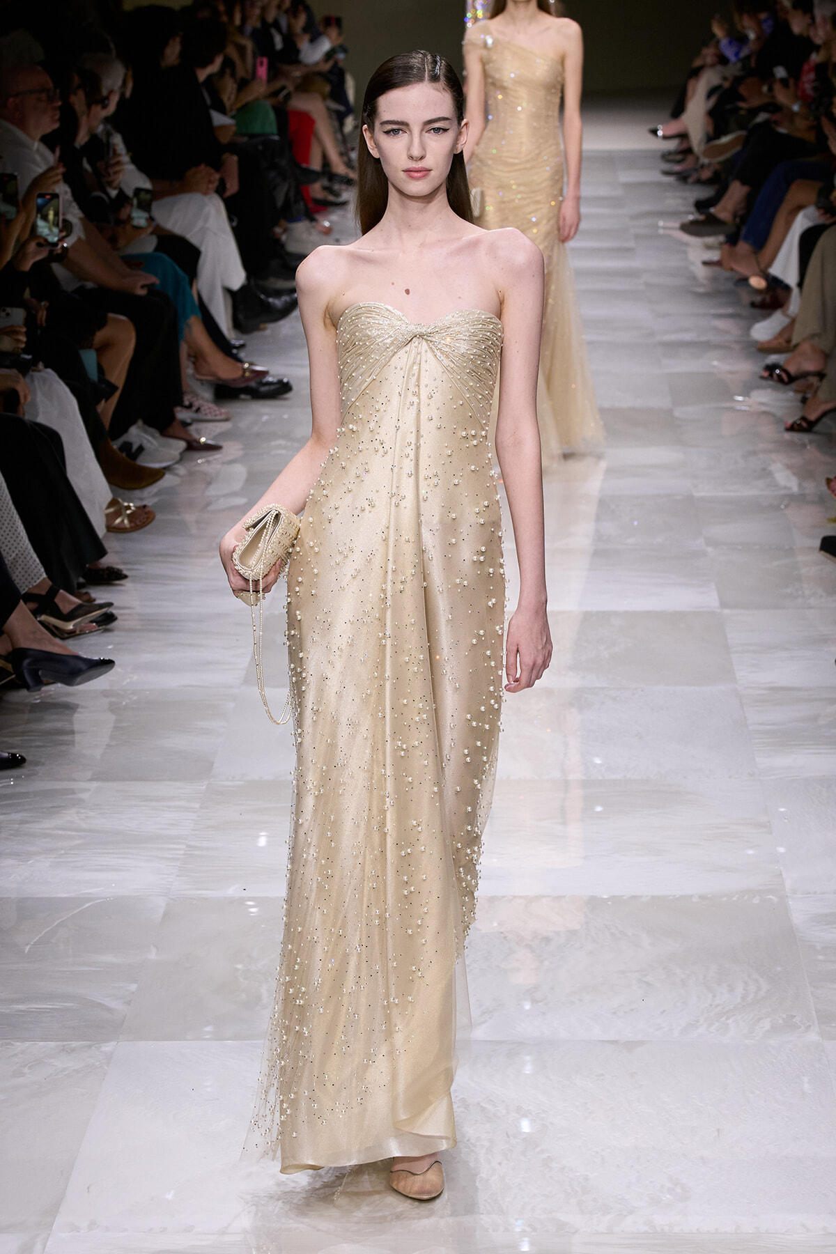 Model on a runway wearing a strapless champagne gown adorned with pearls, holding a small beaded clutch.