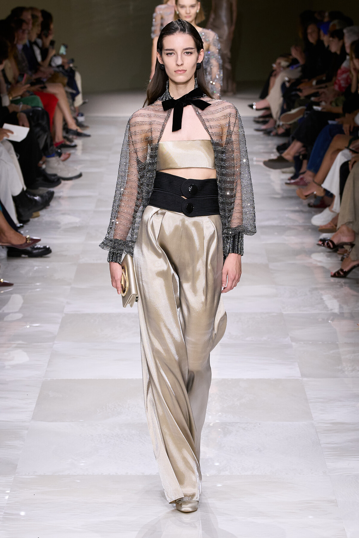 Model walks a runway in champagne satin wide-leg pants, gold tube top, sheer beaded cape, black belt, and bow-neck accessory, clutch in hand