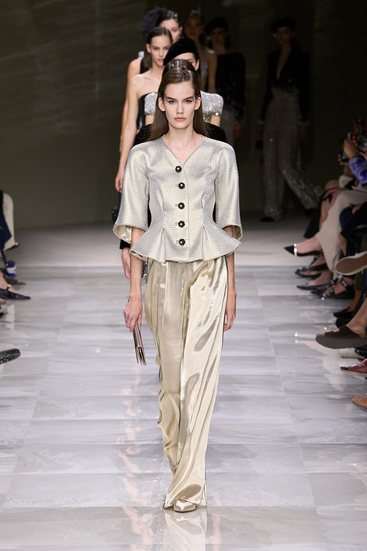 Model walks the runway in a silver textured peplum top with black buttons and cream satin wide-leg pants, holding a clutch.