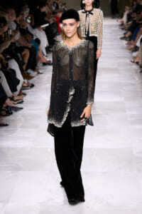 Model on a runway wearing a sheer black beaded top with silver embellishments and wide velvet pants, paired with a black beret.