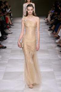 Model walking a runway in a champagne one-shoulder, glittery gown, holding a beige clutch.