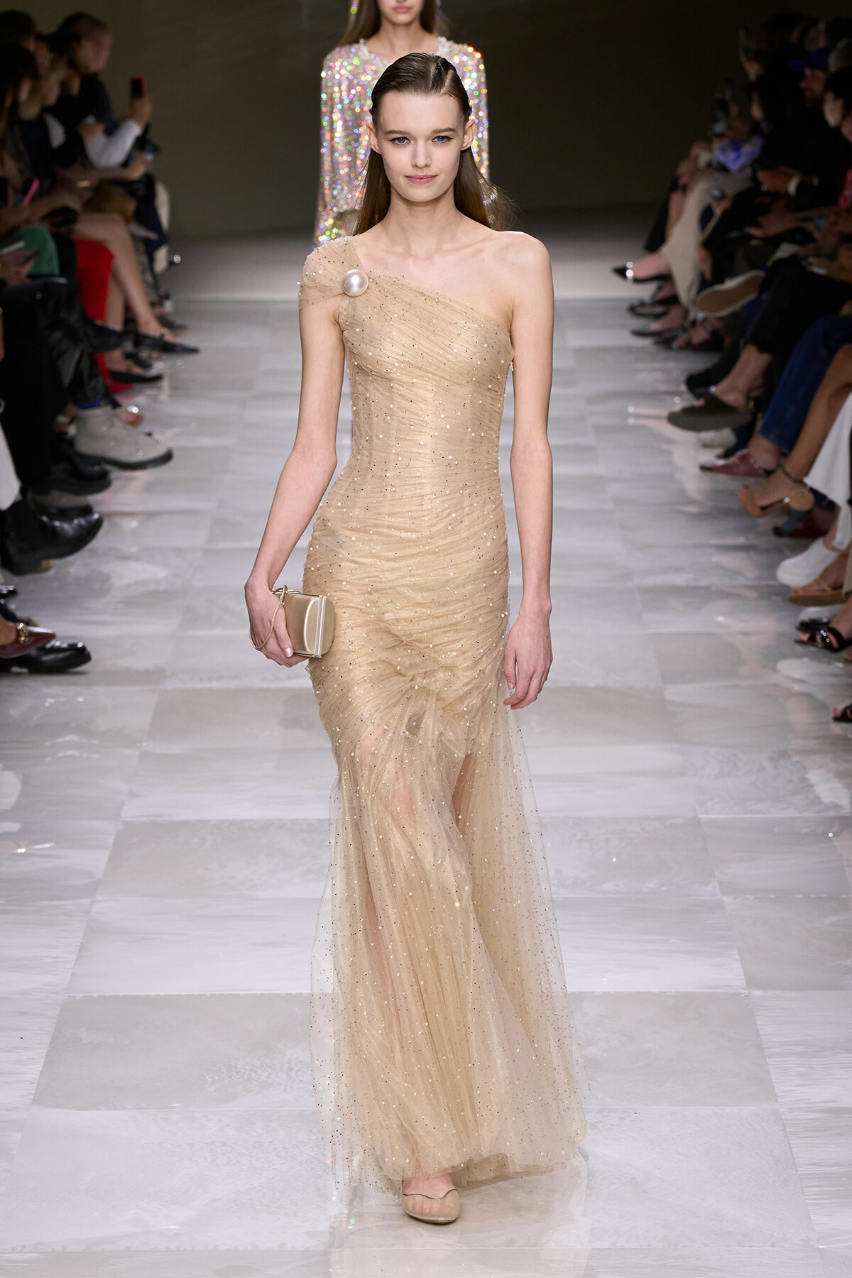 Model walking a runway in a champagne one-shoulder, glittery gown, holding a beige clutch.