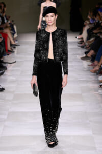 Model on a fashion runway wearing a black beaded cropped jacket and velvet pants with pearl accents, holding a studded clutch.