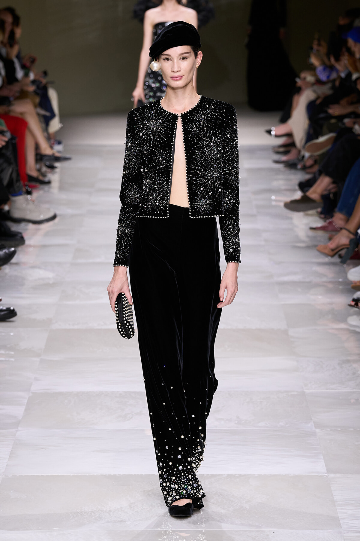 Model on a fashion runway wearing a black beaded cropped jacket and velvet pants with pearl accents, holding a studded clutch.