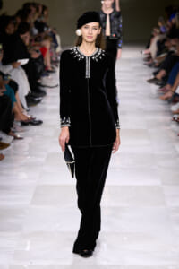Model walks the runway in a black velvet outfit with jeweled collar and cuffs, paired with a matching hat and pearl earrings.