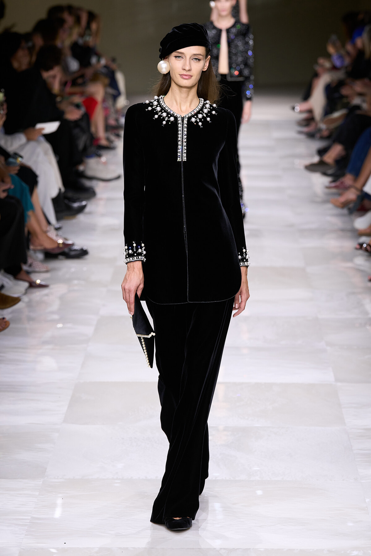 Model walks the runway in a black velvet outfit with jeweled collar and cuffs, paired with a matching hat and pearl earrings.