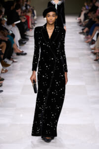 Model walking a runway in a long black velvet gown adorned with pearl-like stars, paired with a black turban and oversized pearl earring.