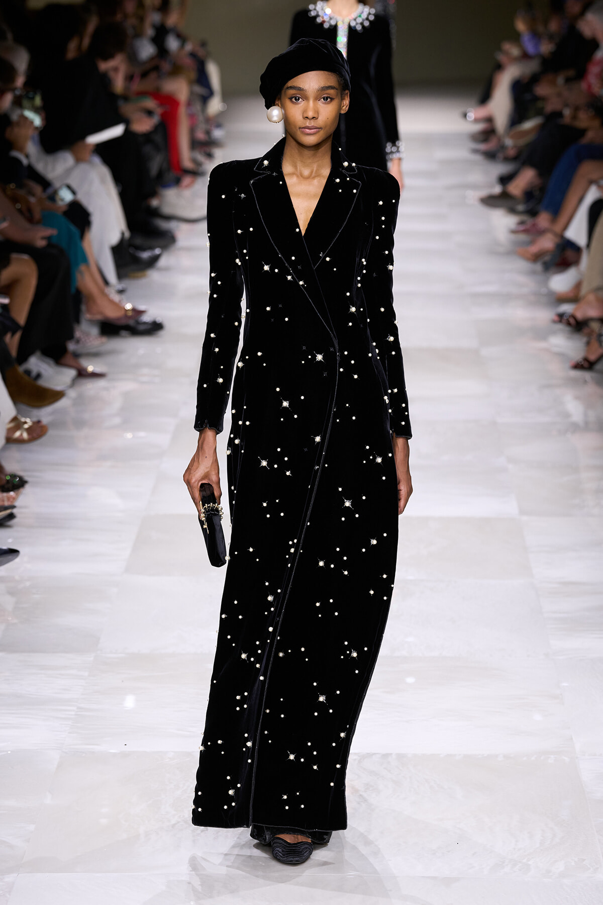 Model walking a runway in a long black velvet gown adorned with pearl-like stars, paired with a black turban and oversized pearl earring.