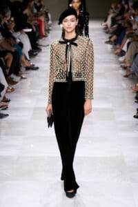 Model walks runway in a sheer beaded jacket with pearl accents over a black velvet jumpsuit, beret, and black accessories.