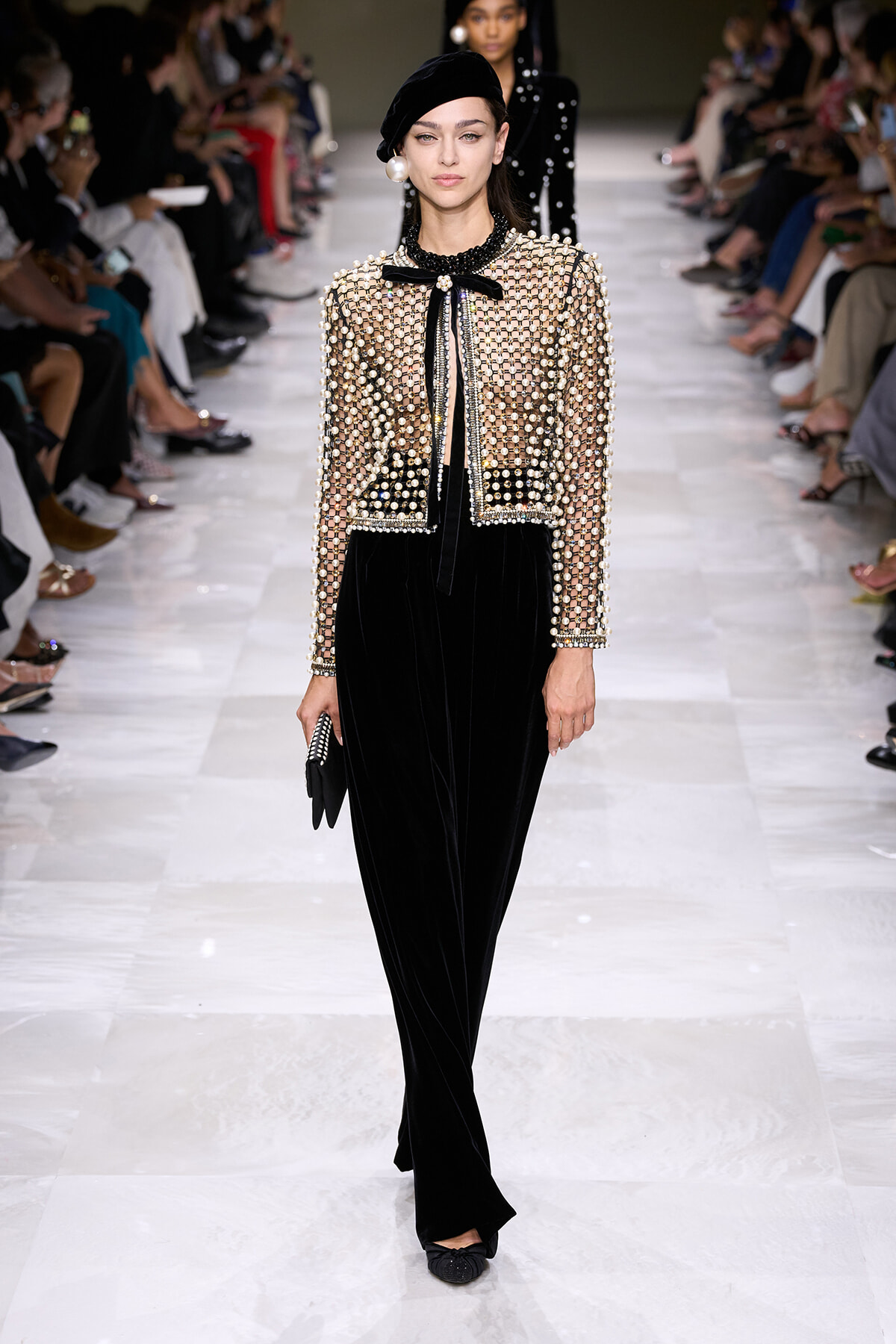 Model walks runway in a sheer beaded jacket with pearl accents over a black velvet jumpsuit, beret, and black accessories.