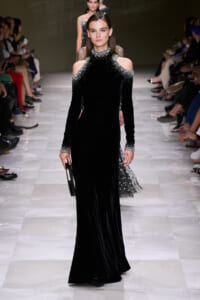 Model walking a runway in a floor-length black velvet gown with crystal-embellished cold-shoulder neckline and cuffs, carrying a small clutch.