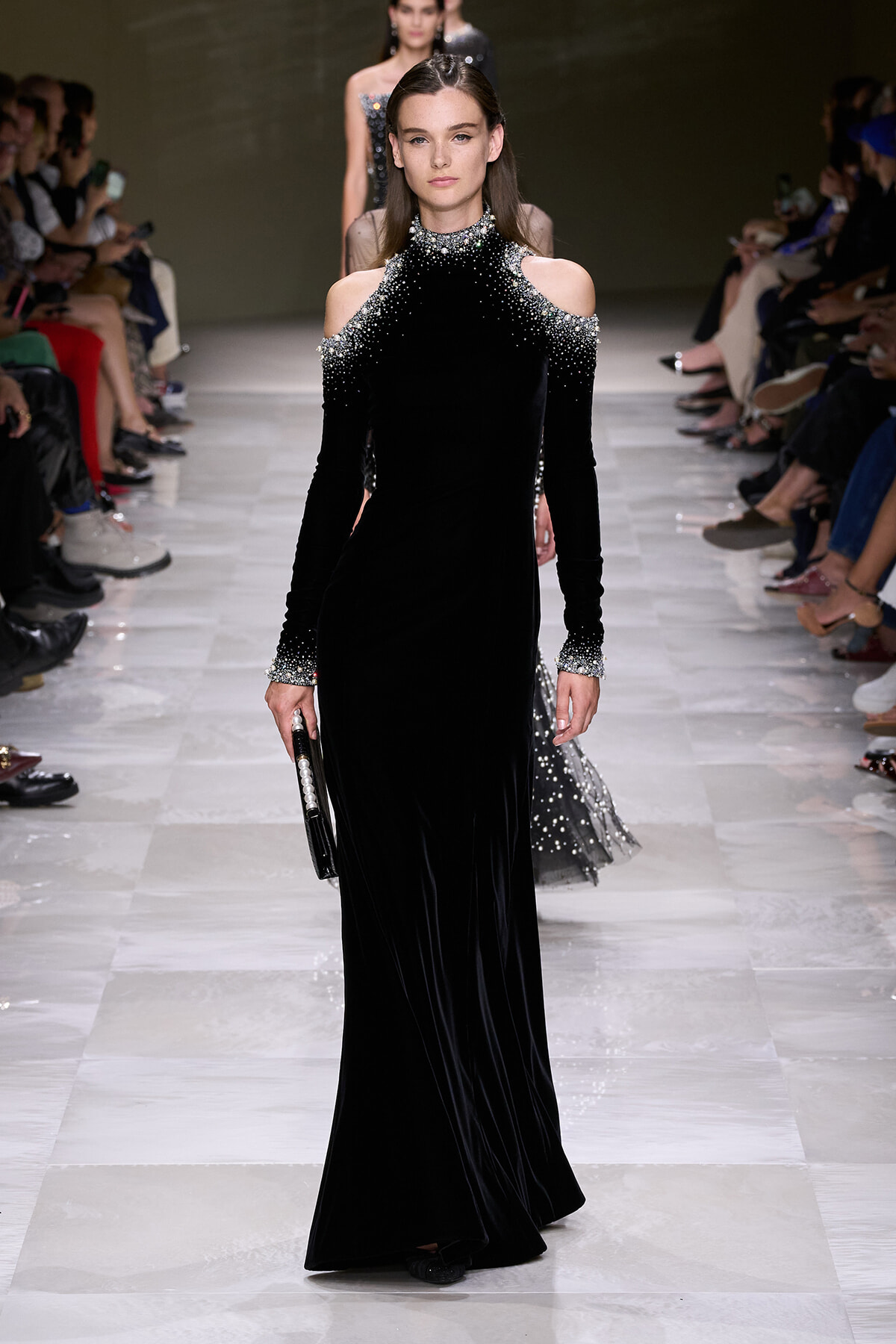 Model walking a runway in a floor-length black velvet gown with crystal-embellished cold-shoulder neckline and cuffs, carrying a small clutch.