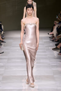 Model on a fashion runway wearing a blush satin strapless gown with crystal-embellished neckline and a beaded clutch in hand.