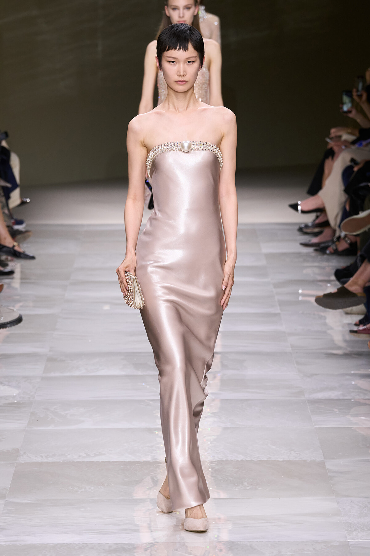 Model on a fashion runway wearing a blush satin strapless gown with crystal-embellished neckline and a beaded clutch in hand.