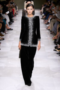 Model walking a runway in a black velvet suit with pearl bead embellishments along the neckline, front, cuffs, and hem; wide-leg pants.
