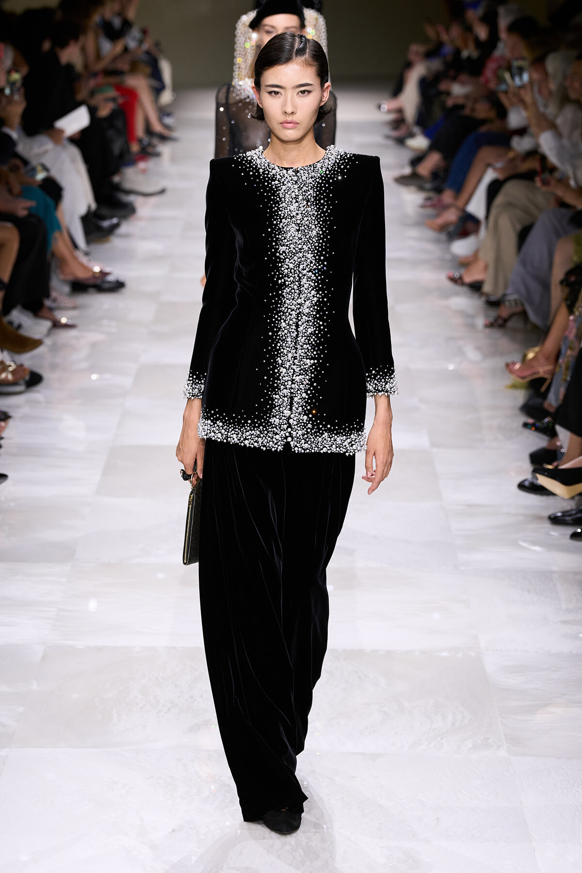 Model walking a runway in a black velvet suit with pearl bead embellishments along the neckline, front, cuffs, and hem; wide-leg pants.