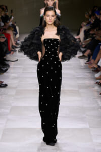 Model walks the runway in a black velvet strapless gown adorned with pearl studs and a voluminous ruffled black cape.
