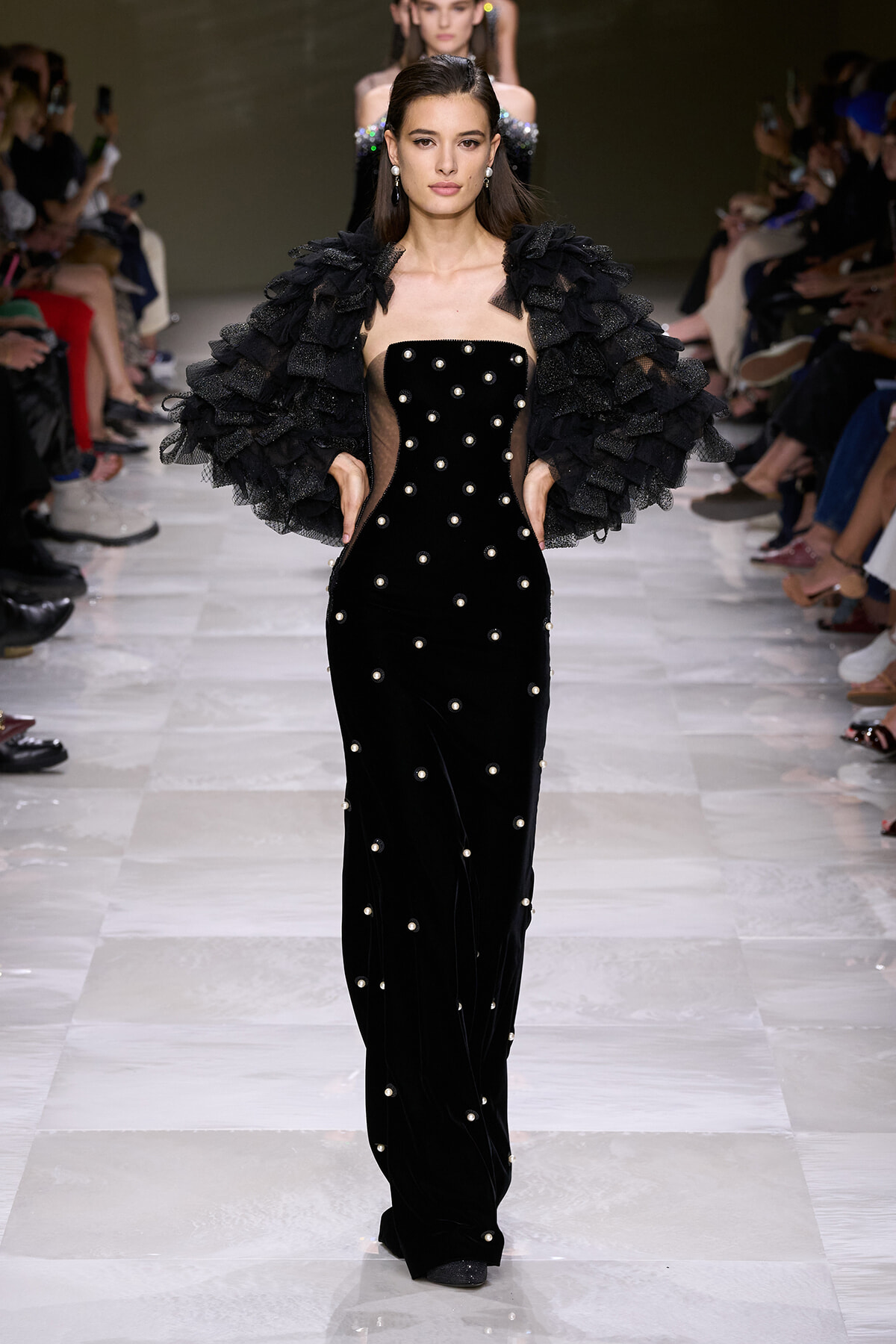 Model walks the runway in a black velvet strapless gown adorned with pearl studs and a voluminous ruffled black cape.