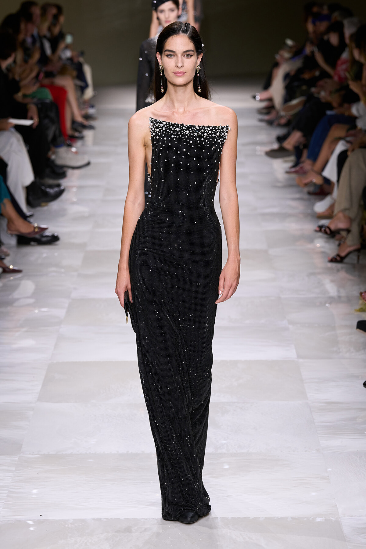 Model walks the runway in a black off-shoulder sequin gown with crystal embellishments, audience on both sides and other models in the background.