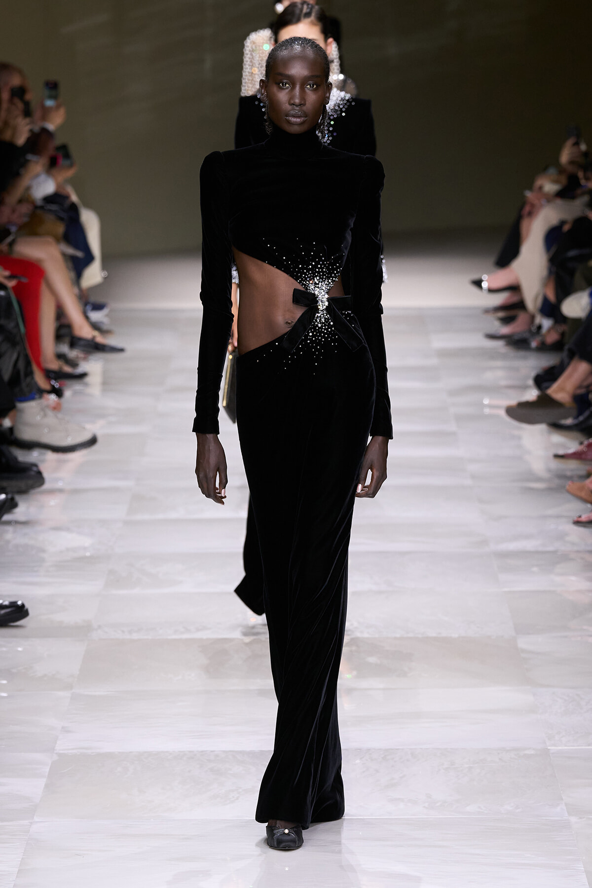 Model on a runway wearing a black velvet cutout gown with sparkling bead details at the waist during a fashion show on a glossy catwalk.