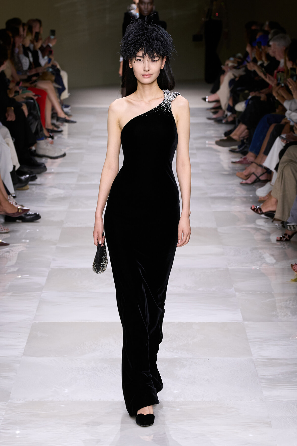 Model walks the runway in a black one-shoulder velvet gown with crystal embellishments at the shoulder and a dramatic black feathered headpiece.