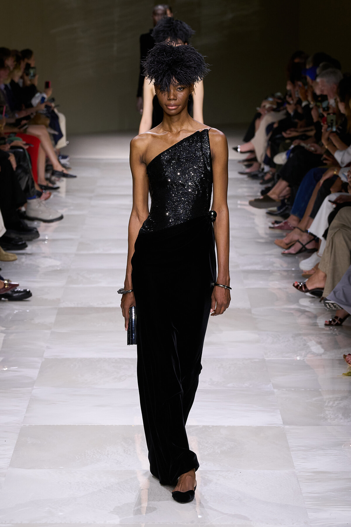 Model walks the runway in a one-shoulder black sequin top and velvet wide-leg pants, with a feathered hat and small clutch.