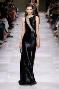 Model walking the runway in a floor-length black sequin gown with a silver-beaded diagonal sash and drop earrings.