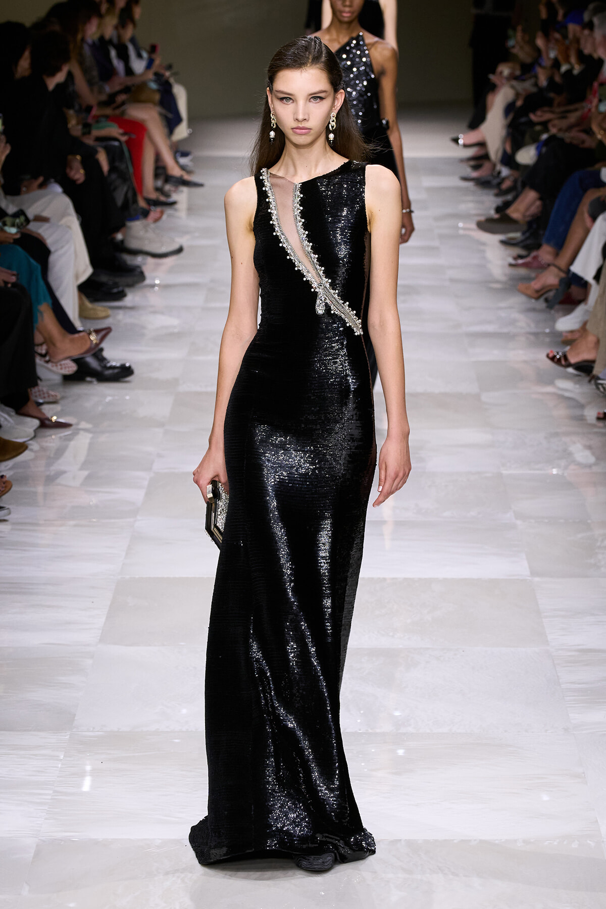 Model walking the runway in a floor-length black sequin gown with a silver-beaded diagonal sash and drop earrings.