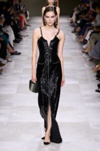 Model on a runway wearing a black sequined evening gown with a V-neck and beaded trim, carrying a studded black clutch.