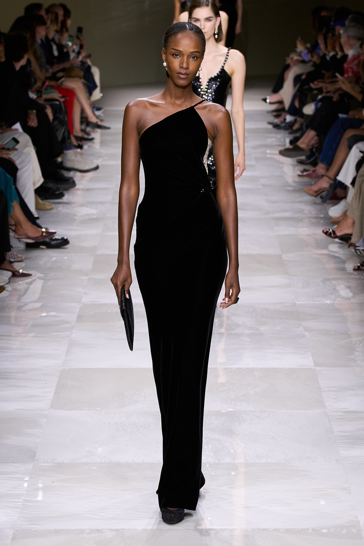 Model walking a fashion runway in a black one-shoulder velvet gown, carrying a black clutch.