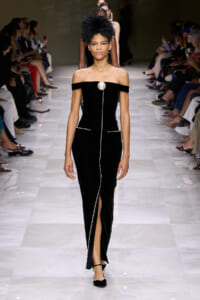 Model walks the runway in an off-shoulder black velvet gown with a large pearl brooch and a vertical beaded seam down the front, finishing in a slit.
