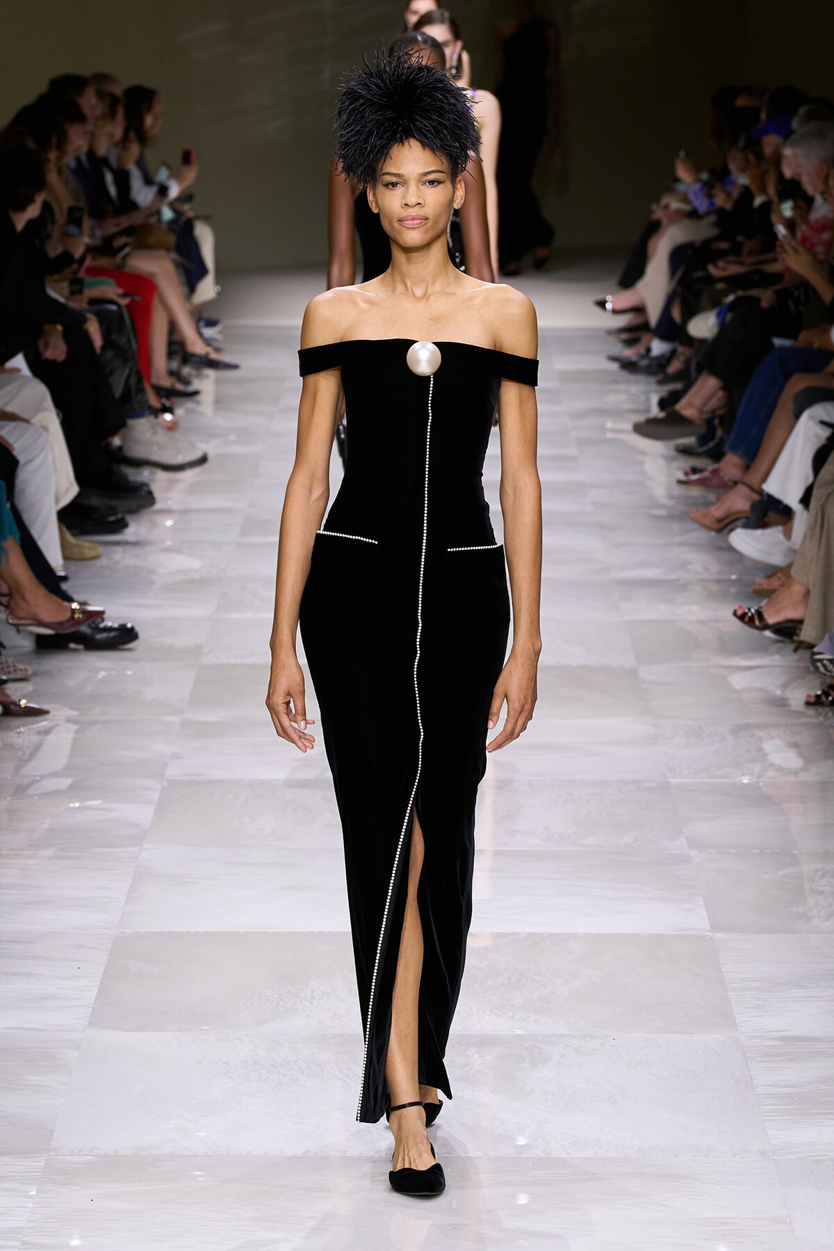 Model walks the runway in an off-shoulder black velvet gown with a large pearl brooch and a vertical beaded seam down the front, finishing in a slit.