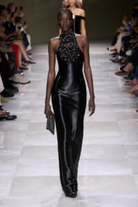Model on a runway wearing a black satin halter gown with a heavy beaded neckpiece, holding a small striped clutch.