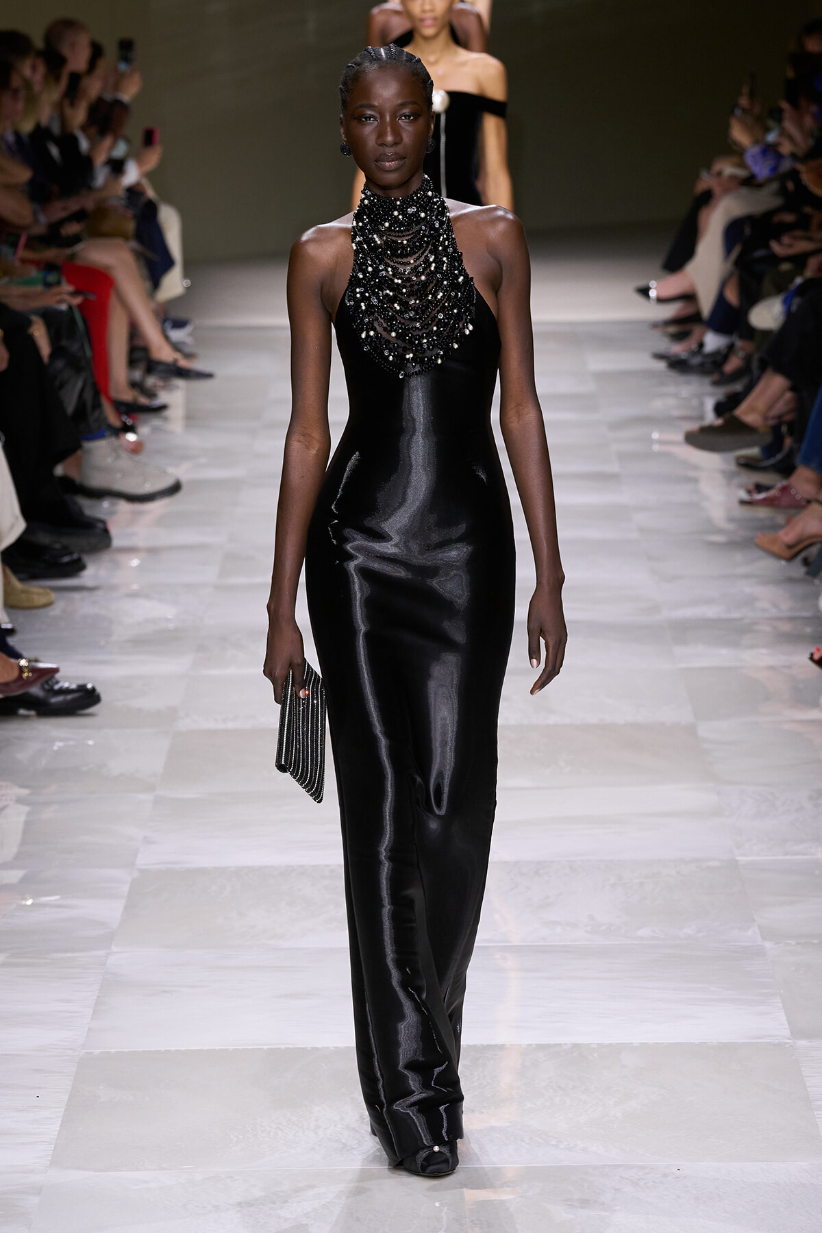 Model on a runway wearing a black satin halter gown with a heavy beaded neckpiece, holding a small striped clutch.