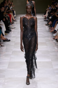 Model walking the runway in a sheer black sequin gown, with delicate straps and a form-fitting silhouette, at a fashion show.