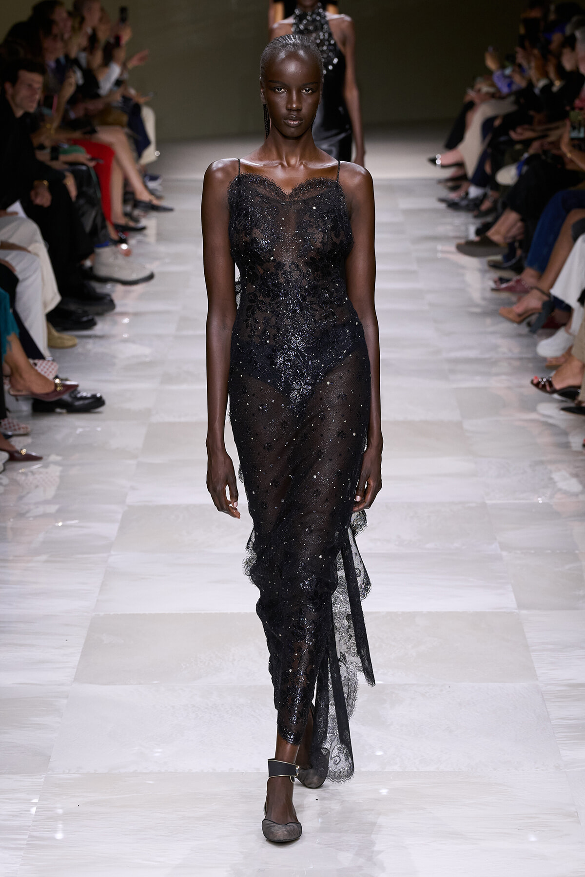 Model walking the runway in a sheer black sequin gown, with delicate straps and a form-fitting silhouette, at a fashion show.
