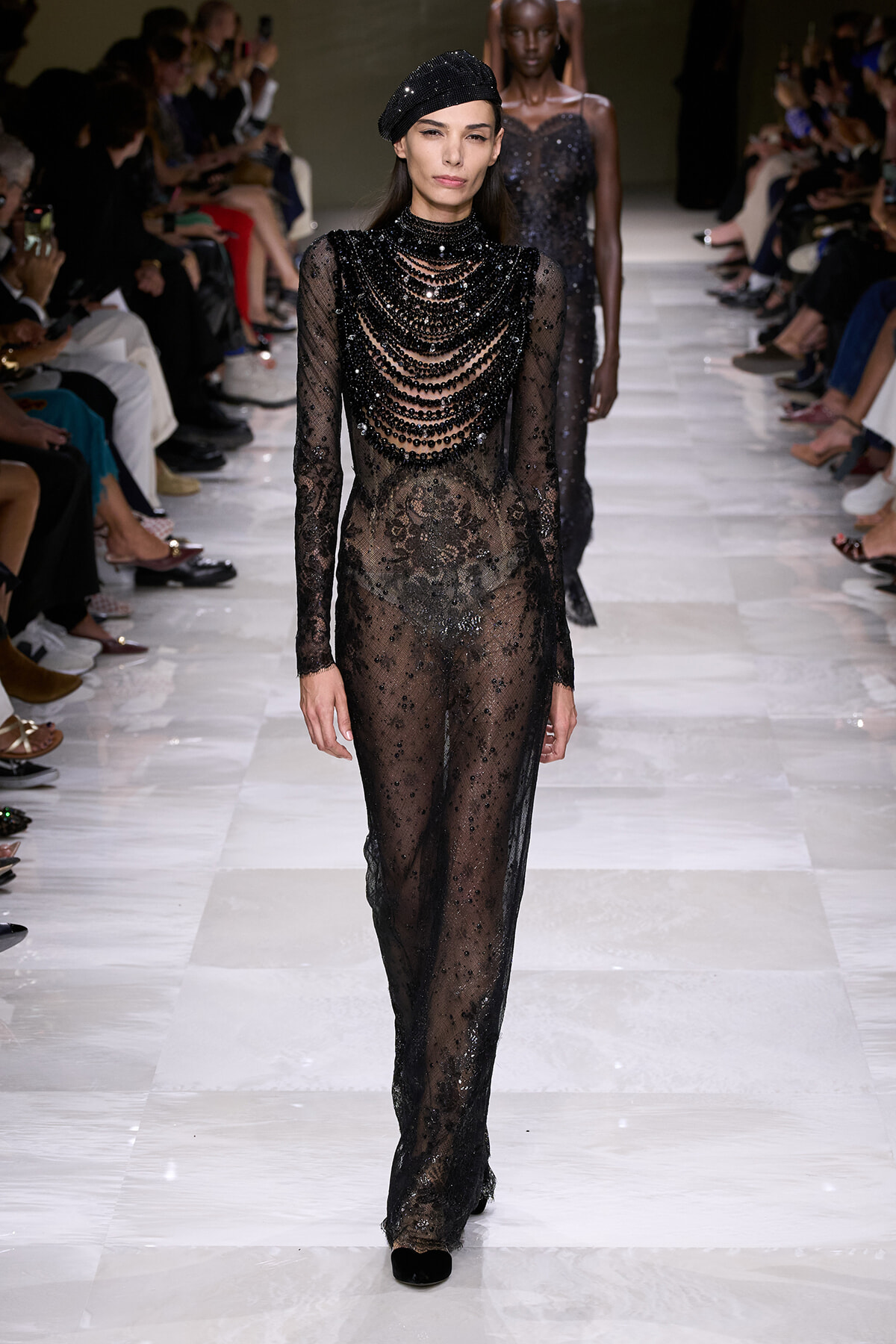 Model walks the runway in a sheer black lace gown with layered beaded necklaces and a black beret, audience seated along the sides.